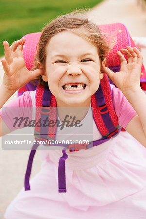 Portrait of 5 year old schoolgirl with her pink and purple school bag, making faces and looking at camera, showing her front teeth missing