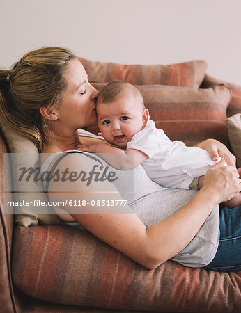 A woman lying on a sofa playing with a baby girl, kissing her head.