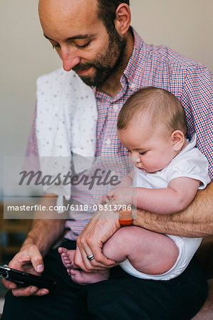 A man holding his small child in the crook of his arm, texting on his smart phone with the other.