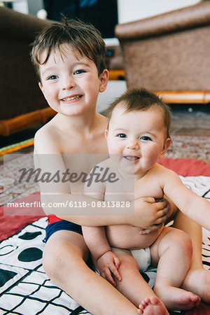 Two siblings, a boy and his baby sister sitting on a rug laughing.