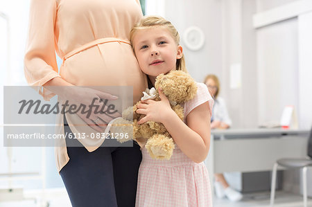 Portrait girl with teddy bear hugging pregnant mother in doctor's office