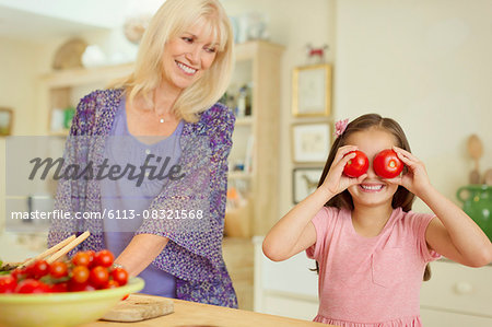 Portrait playful granddaughter covering eyes with tomatoes in kitchen