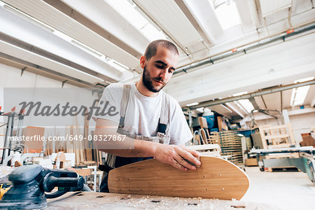 Young man in carpentry workshop looking down sanding skateboard