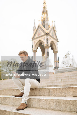 Young man sitting on steps reading smartphone texts, Albert Memorial, London, England, UK