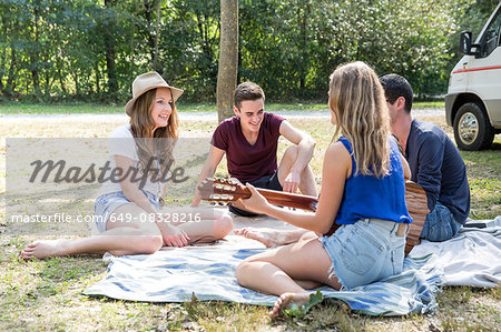 Group of young adults sitting on picnic blanket , young woman playing guitar