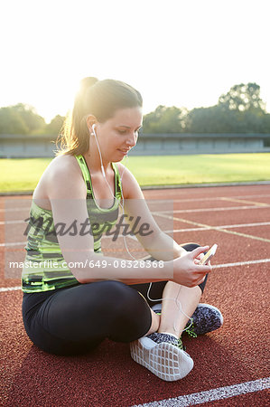 Young female runner cross legged on race track reading smartphone texts