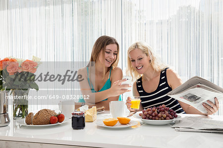 Women sitting at dining table enjoying a continental breakfast together, looking at cellular phone smiling