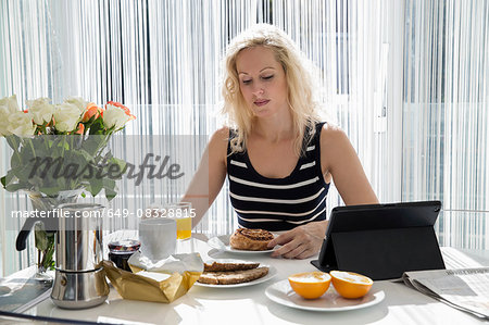 Mature woman sitting at table with digital tablet looking down eating breakfast