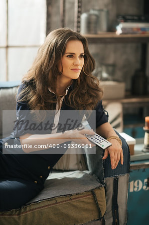 Stylish elegant woman relaxing on sofa and watching tv in loft apartment. Urban chic loft decoration details and window. Modern lifestyle concept.