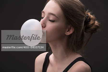 Portrait of brown-haired, which inflates languidly pink bubble of chewing gum. Photographed in studio on a dark background.
