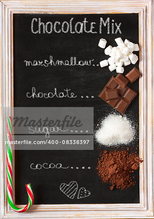 Hot chocolate mix on old chalk blackboard. Sweet marshmallow, chocolate bars, sugar, cocoa powder and mint candy cane for cooking winter holiday drink.