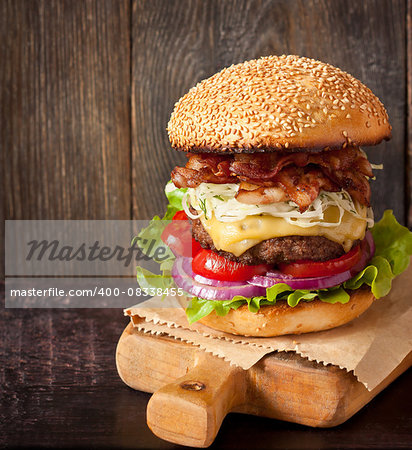 Big delicious cheeseburger stacked high with a bacon, beef patty, coleslaw, cheese, lettuce, red onion and tomato on sesame seed bun served on wooden cutting board.