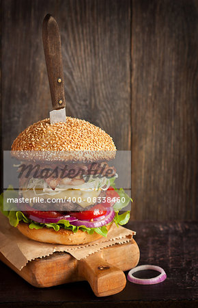 Big delicious cheeseburger stacked high with a bacon, beef patty, coleslaw, cheese, lettuce, red onion and tomato on sesame seed bun served old knife on wooden cutting board. Rustic style.