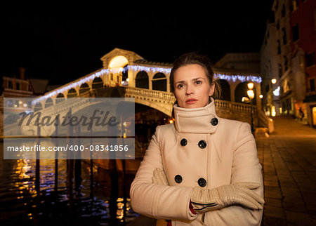 Pensive elegant young woman in white coat standing in front of Rialto Bridge in the evening. She having Christmas time trip and enjoying stunning views of Venice, Italy