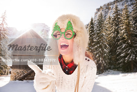 Going wild for Christmas season in a secluded spot in the countryside. Smiling young woman in funny Christmas tree glasses pointing on something in the front of a cosy mountain house.