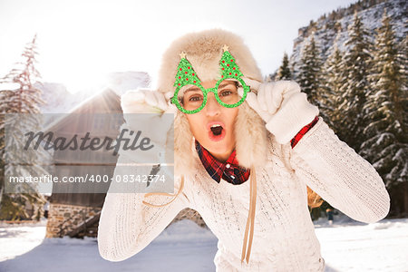 Going wild for Christmas season in a secluded spot in the countryside. Portrait of merry young woman in funny Christmas tree glasses having fun in the front of a cosy mountain house.