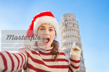 Young, itching from energy and searching for excitement. I'm going to Christmas trip to Italy. It is a no-brainer. Happy woman in Santa hat taking selfie and pointing on Leaning Tour of Pisa.