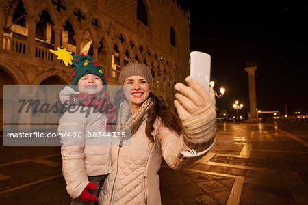 Holiday family trip to Venice, Italy can change the whole Christmas experience. Happy mother with child taking selfie with smartphone while standing on Piazza San Marco in the evening. Winter Tourism