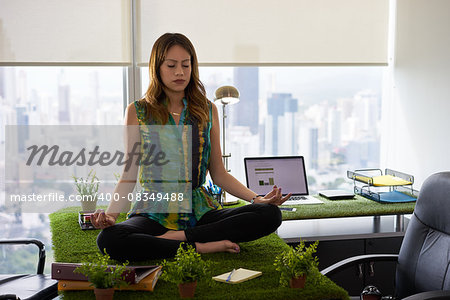 Young hispanic woman in office, sitting on desk covered with grass and plants. The businesswoman does zen and yoga meditation in lotus position. Full length, front view