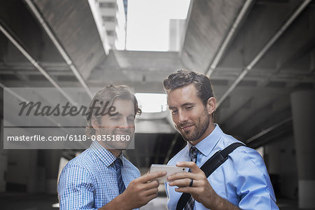 Two men in shirts and ties taking a selfie with a smart phone, with an urban walkway and tall buildings in the background.