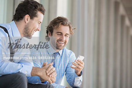 Two business men in shirts and ties sitting outside an office building, checking their smart phones.