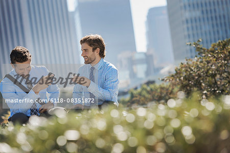 Two men in shirts and ties in a park in the city holding their smart phones.