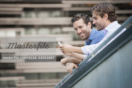 Two men leaning on a railing, one holding a smart phone.