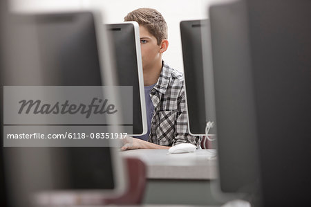 A school room, computer laboratory or lab with rows of computer monitors and seating. A young person seated working at a terminal.
