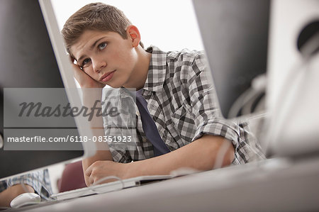 A school room, computer laboratory or lab with rows of computer monitors and seating. A boy sitting with his hand on his chin looking fed up.
