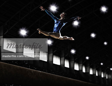 A female gymnast, a young woman performing a floor routine, in mid air with legs and arms outstretched and back bent.