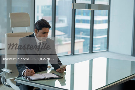 Businessman using mobile phone and taking notes at desk