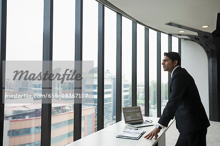 India, Businessman standing by desk and looking through window in office