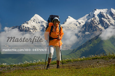 Woman (backpacker)  with sunglasses and sticks against mountains covered with snow background. Tourist is situated on alpine meadow. Tviber mount is in the left part of image.