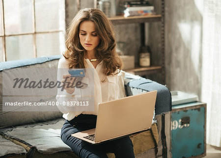 Elegant dressed young woman is sitting on couch and typing info from credit card to laptop