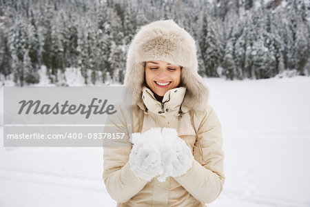Magical mix of winter season and mountain landscape create the perfect mood. Happy young woman in white coat and fur hat looking on snow in the hands while standing outdoors