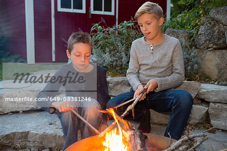 Two boys with sticks sitting by garden campfire at dusk