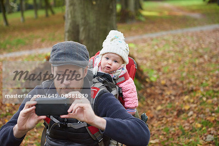 Mid adult man in park wearing flat cap carrying daughter on back in baby carrier taking selfie using smartphone