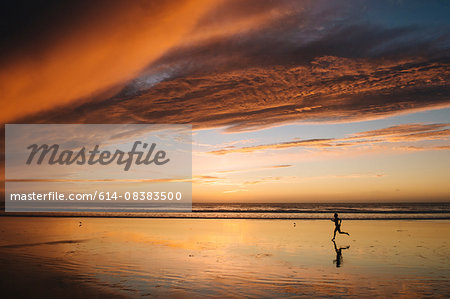 Side view of mid adult nude womans silhouette running on beach under dramatic sky at sunset