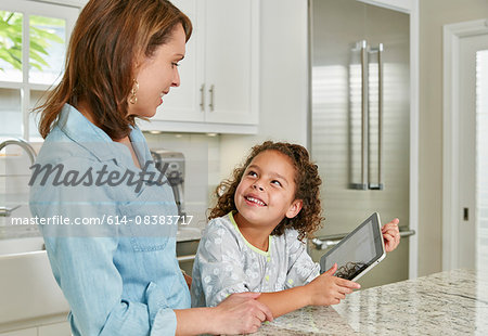 Mother and daughter at kitchen counter using digital tablet, looking up smiling