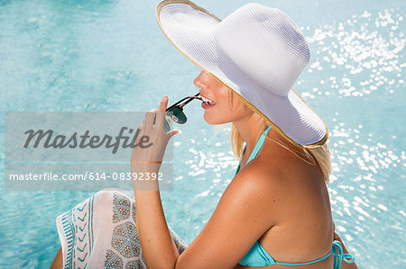 High angle side view of young woman wearing sunhat sitting by swimming pool holding sunglasses
