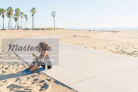 Mid adult woman sitting by beach, using smartphone
