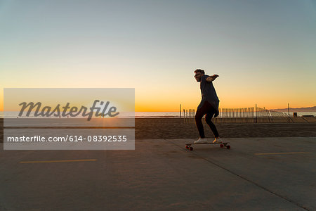 Young man using skateboard on pathway, beside beach, sunset