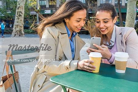 Young adult female twins with takeaway coffee reading smartphone texts in city park