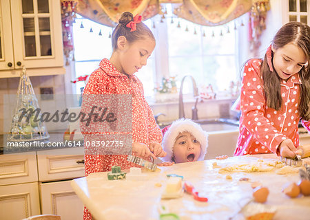 Boy in kitchen wearing santa hat making cookies with sisters peeking over kitchen counter open mouthed