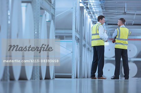 Businessmen in reflective clothing talking near large paper spools in printing plant