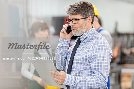 Businessman with clipboard talking on cell phone in factory