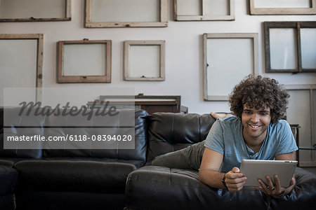 Loft decor. A wall hung with pictures in frames, reversed to show the backs. A man lounging on a sofa, using a digital tablet.