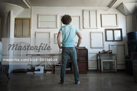 Loft decor. A wall hung with pictures in frames, reversed to show the backs. A man with hands on his hips looking at the wall.