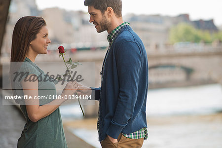 A couple in a romantic setting by a river. A man offering a woman a red rose.