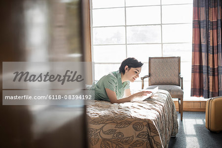 A young woman lying on a bed in a motel room using a digital tablet touchscreen.